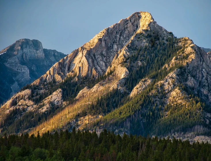 a mountain with trees in the foreground and a blue sky in the background, a picture, by Andrew Domachowski, shutterstock, backlit golden hour, montana, big sharp rock, dappled in evening light