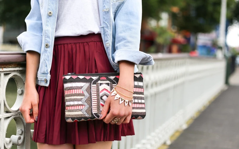 a woman standing on a bridge holding a purse, a picture, pleated skirt, detailed jewelry, istock, aztec street fashion