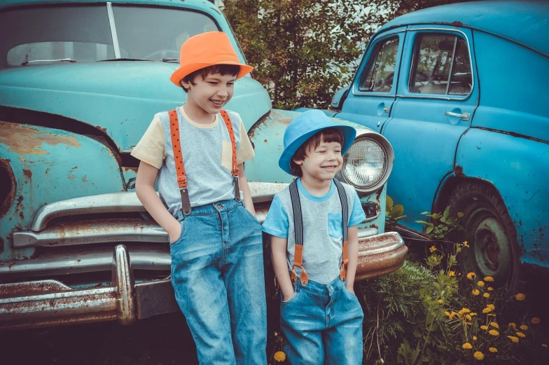 two young boys standing next to an old car, by Joe Bowler, pexels, cute hats, blue!! with orange details, overalls, retro stylised