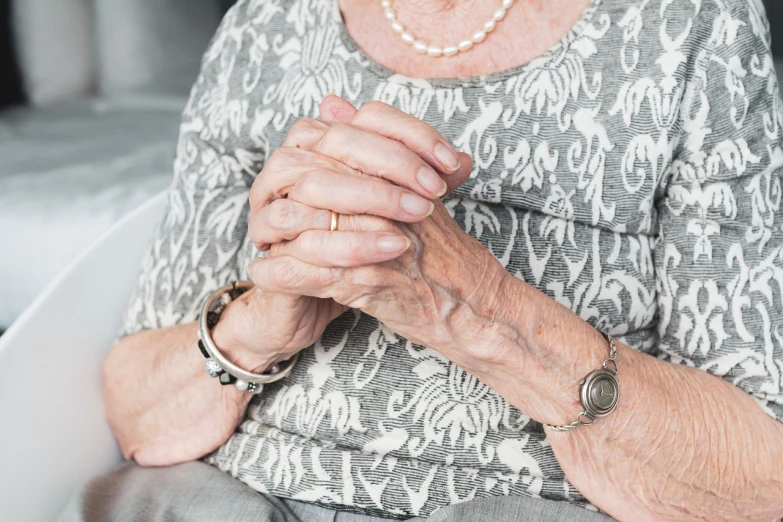 a woman sitting in a chair with her hands folded over her chest, pexels, elderly woman, wearing two silver bracelets!, photo pinterest, disease