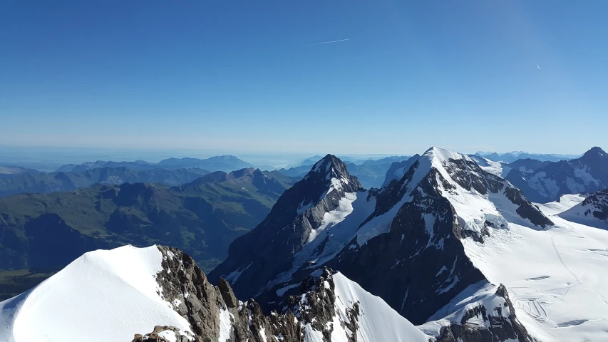 a man standing on top of a snow covered mountain, by Caroline Mytinger, pixabay, in the swiss alps, clear blue skies, viewed from bird's-eye, summer morning