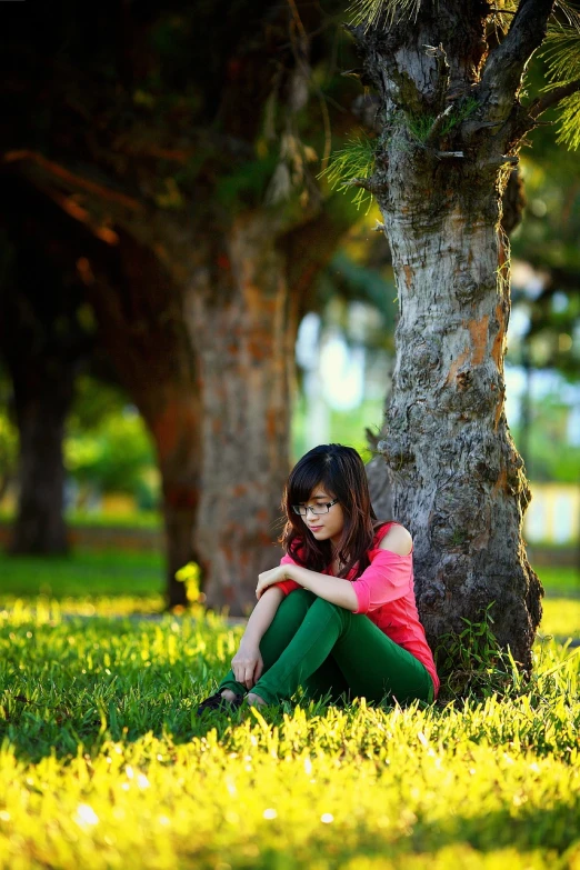 a woman sitting in the grass next to a tree, a picture, by Basuki Abdullah, pixabay, beautiful lonely girl, shades green and red, girl with glasses, 7 0 mm. dramatic lighting