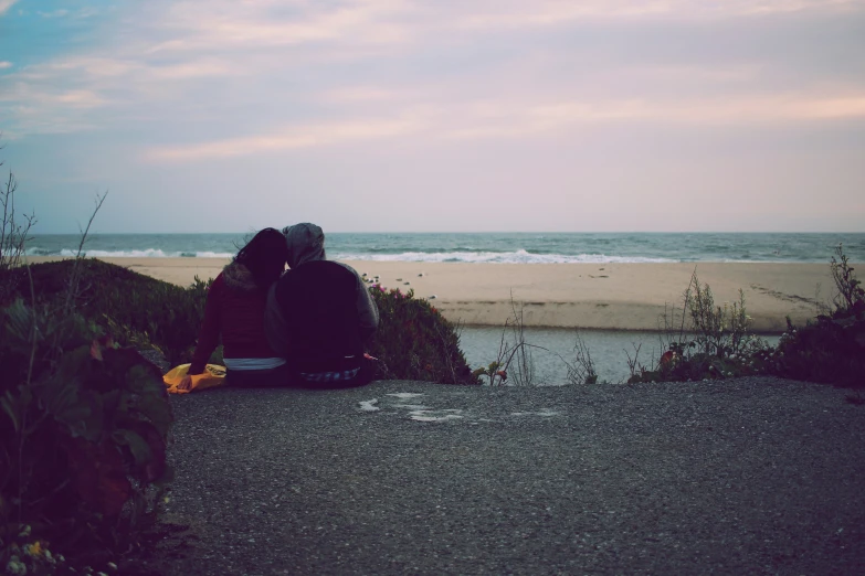 two people sitting on the side of a road watching the ocean