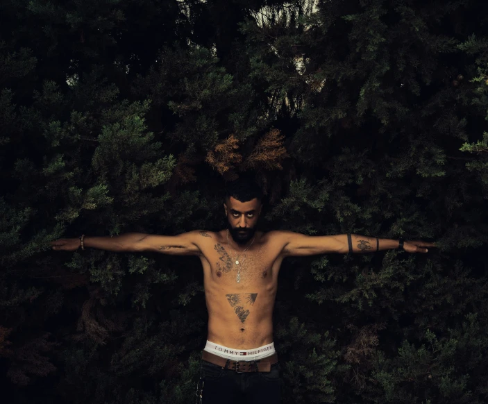 a shirtless man standing in a wooded area, showing off his tattoos