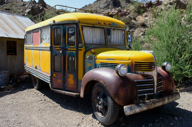 an old yellow school bus sits outside on gravel near some bushes and trees