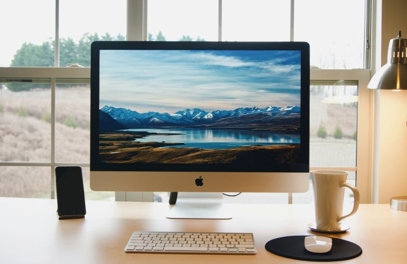 an apple computer sitting on a desk near some windows