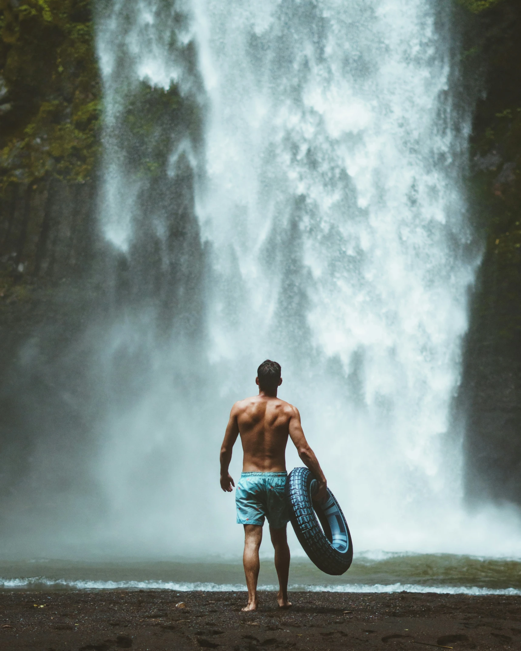 a man standing in front of a waterfall while holding a surfboard