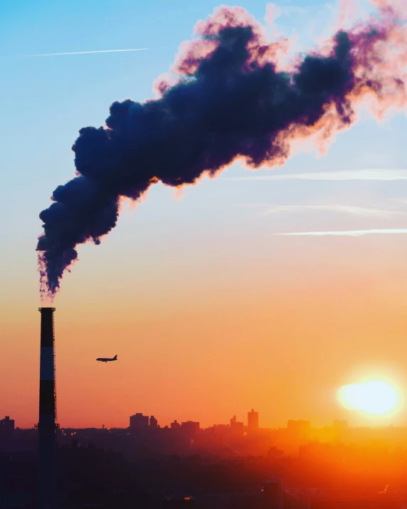 an airplane flying past a smoke stack and setting sun