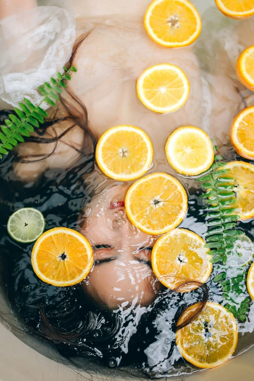 a woman has fallen asleep in a water filled bath with many oranges and ferns