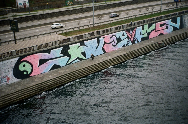 a graffiti - filled wall beside water at the side of a bridge