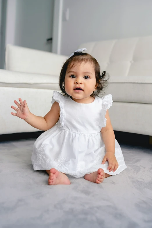 a baby girl in a white dress sitting on the floor