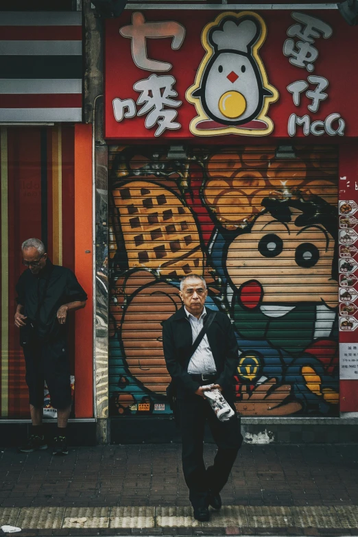 a man wearing black clothes and a suit, leaning against a store door with teddy bears painted on it