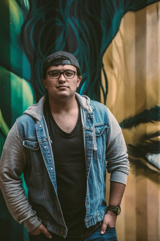 a man in denim vest and hat standing in front of a wall with graffiti