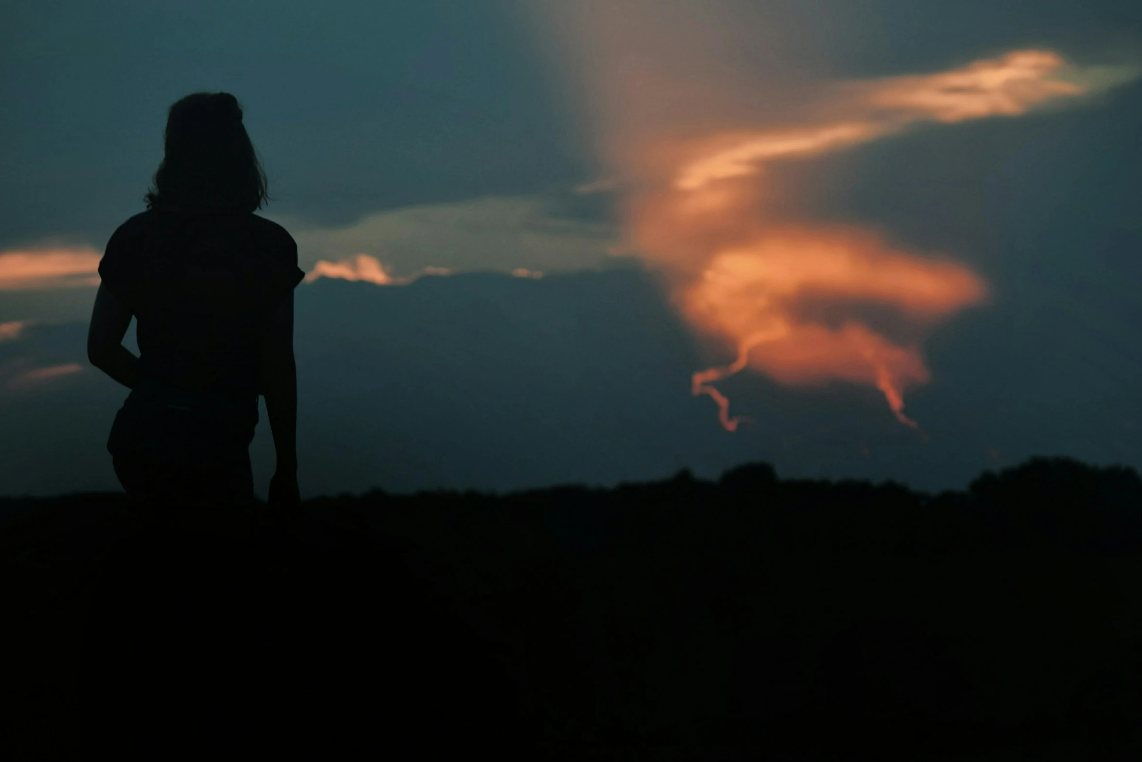 a silhouetted girl with her back turned stands in front of a sunset