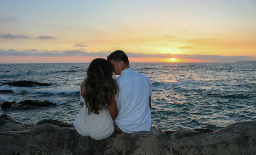 a couple are sitting on a rock as the sun sets in the background