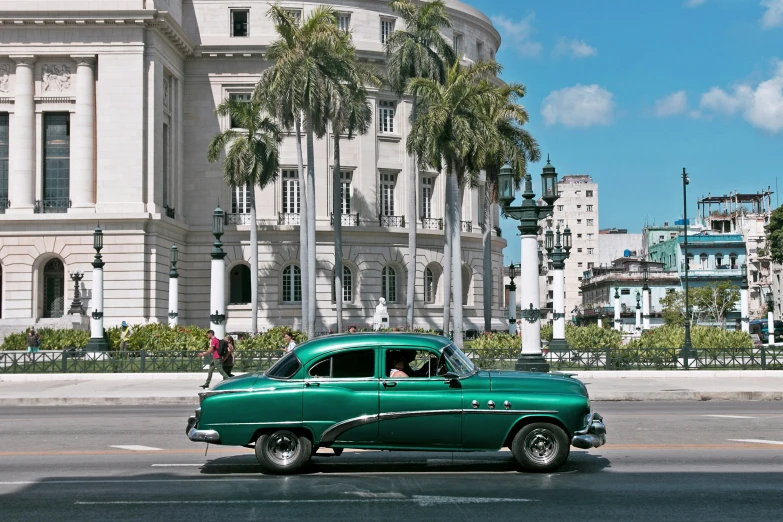 an old green car driving past a large building