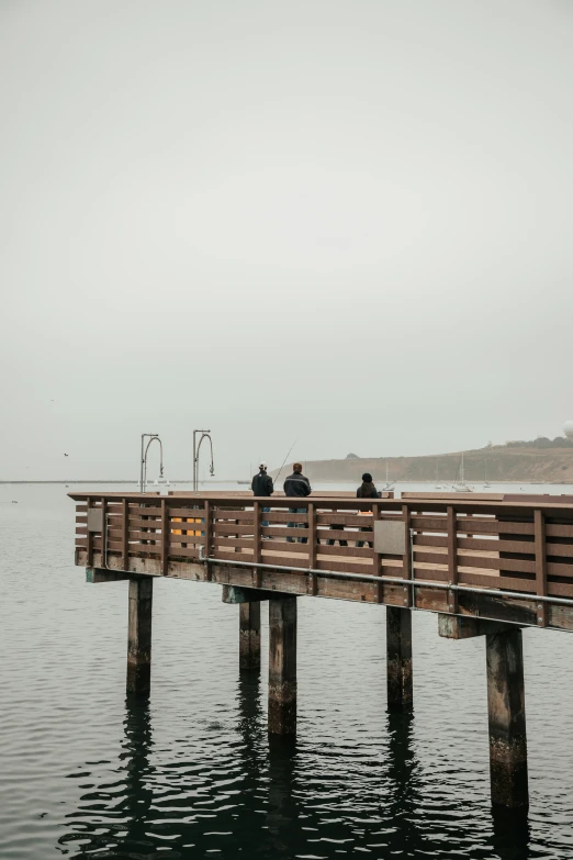 three people sitting on a long wooden dock overlooking the ocean