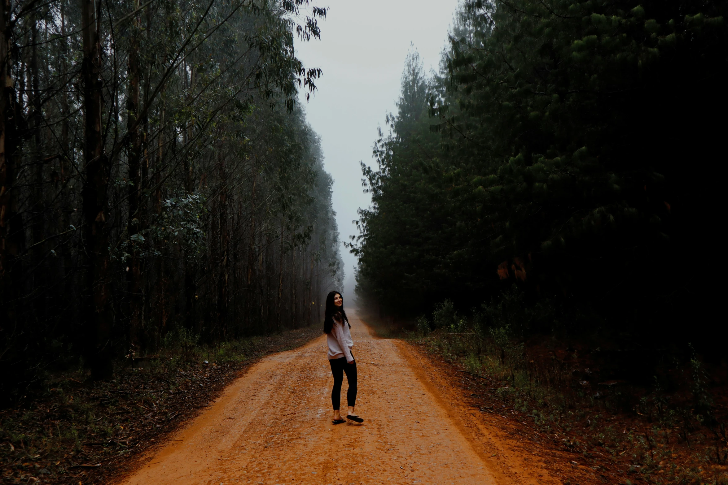 a woman walking across a dirt road near some trees