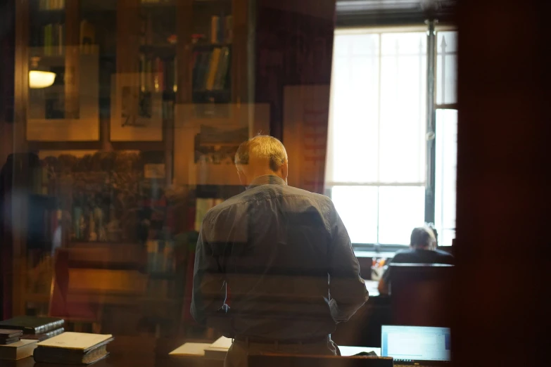 an individual stands with his back turned, looking towards a wall covered with shelves with books