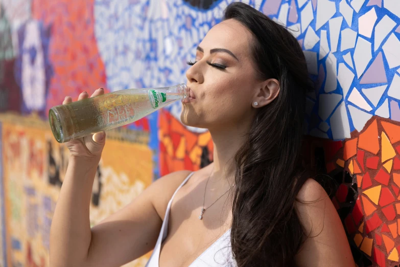 a woman is drinking from a bottle and leaning against a colorful wall