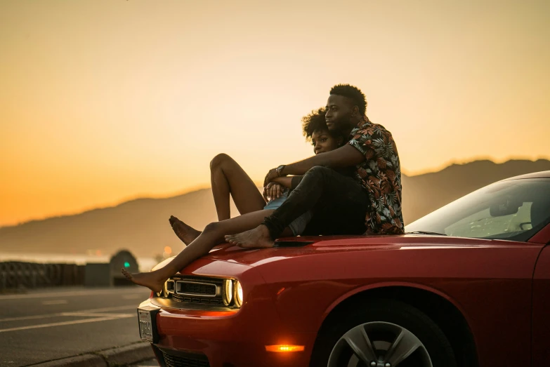 two people sitting on top of a red car near water