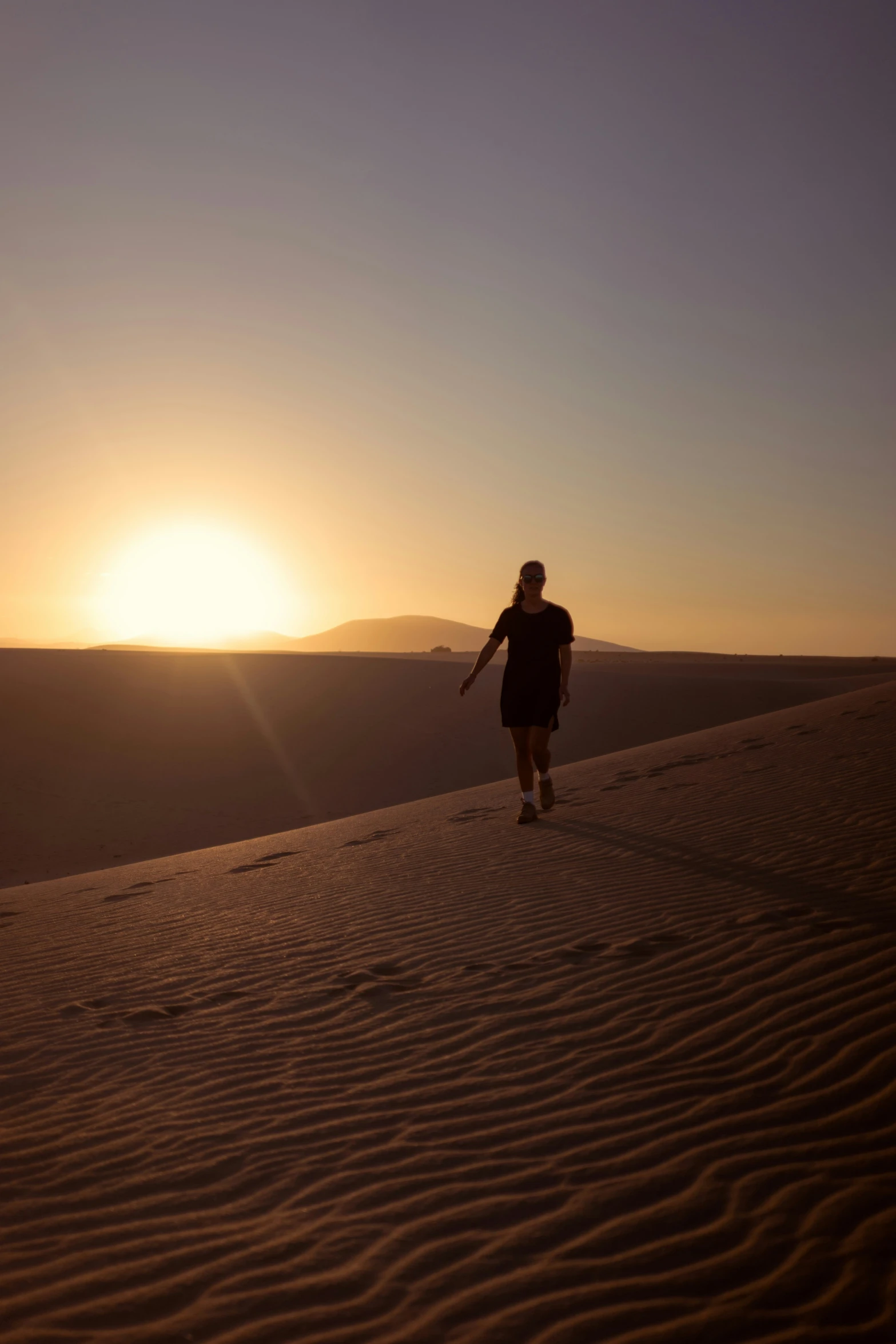 man walks through sand dunes at sunrise with a mountain range in the background