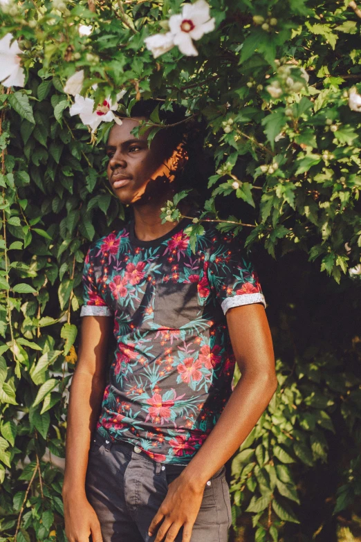 an image of man with flowered t - shirt standing with a green bush and shrub behind him