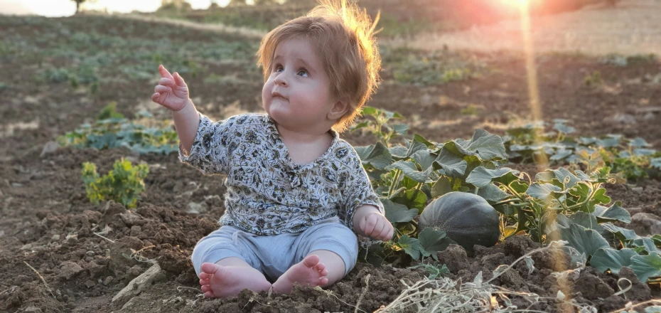 baby girl sits on dirt with large green plants in background