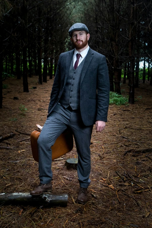 a bearded man is sitting on his luggage in a dark wooded area