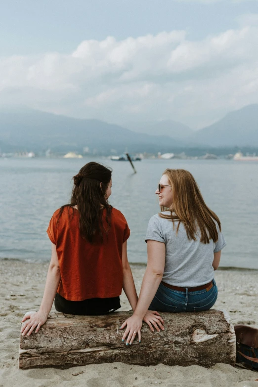 two woman sitting on a log in front of the water