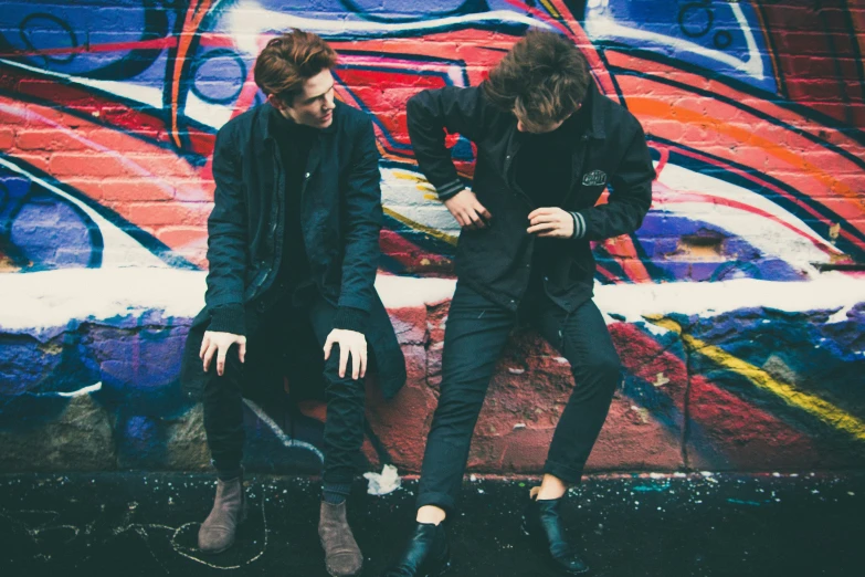 two men in front of a graffiti wall with one on his knees