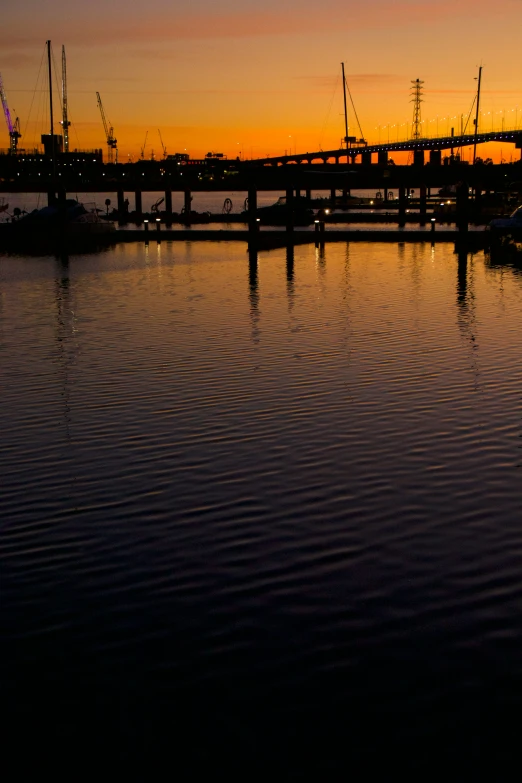 sunset over a bridge with boats and cranes in the background