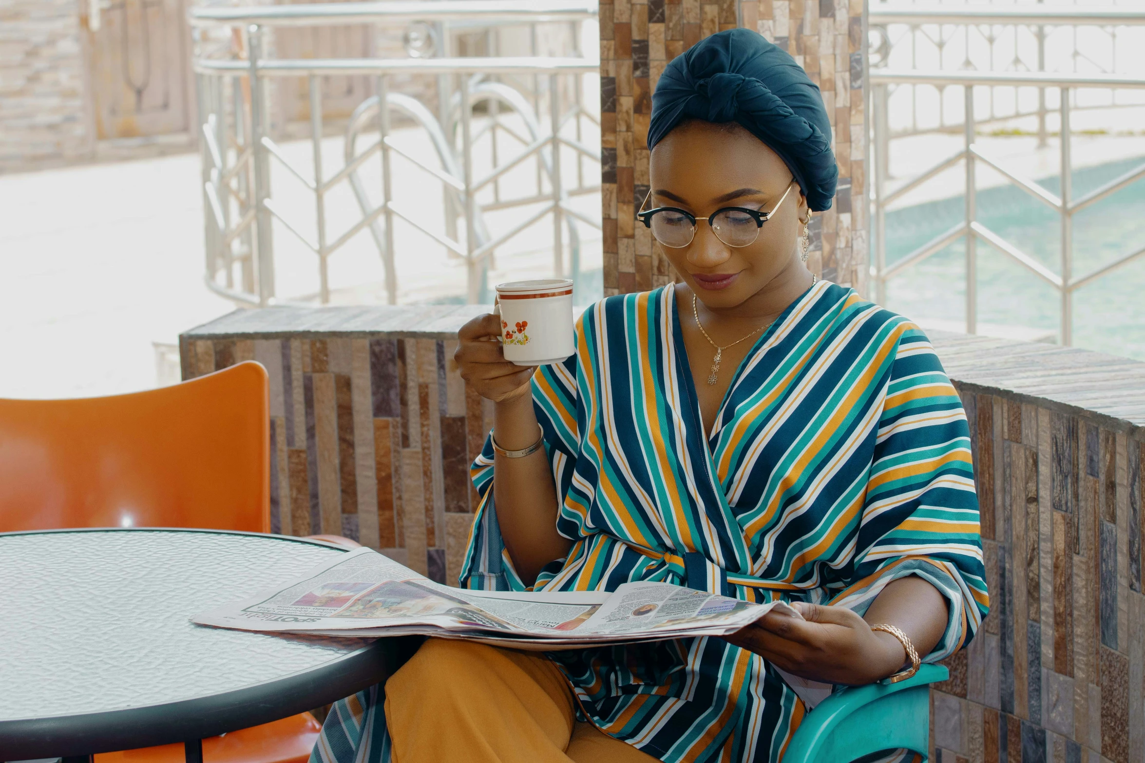 a woman is sitting at an outdoor table reading a magazine and drinking coffee