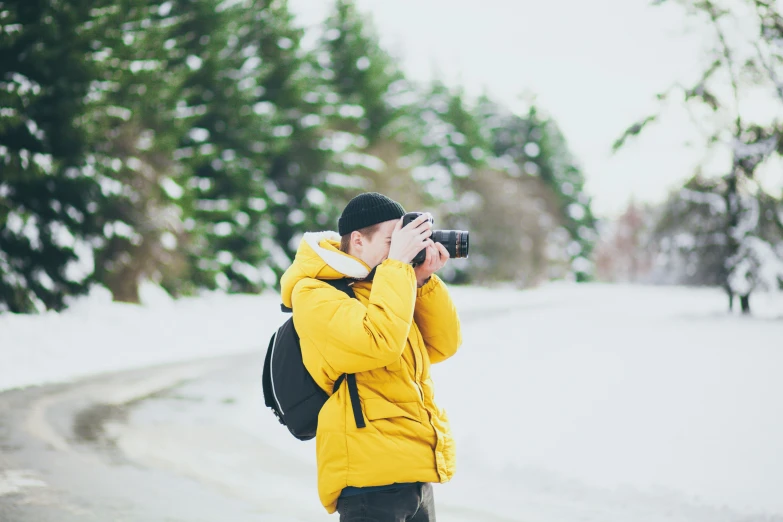 man in yellow jacket taking a po of snow and pine trees