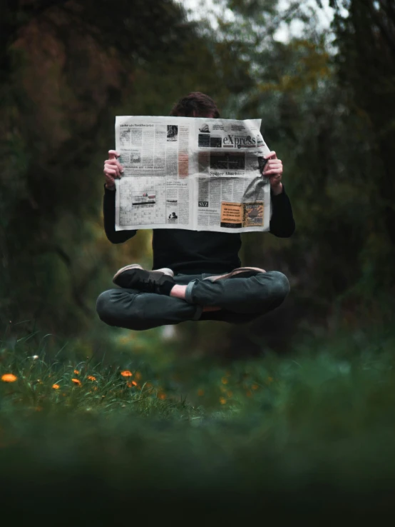 person sitting in grass while holding newspaper with trees and grass behind