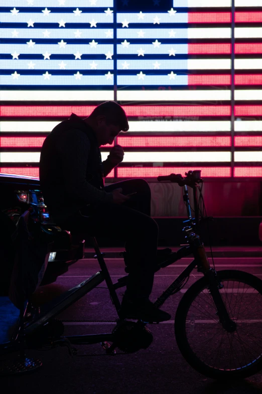 a man sits on a bicycle next to a large american flag