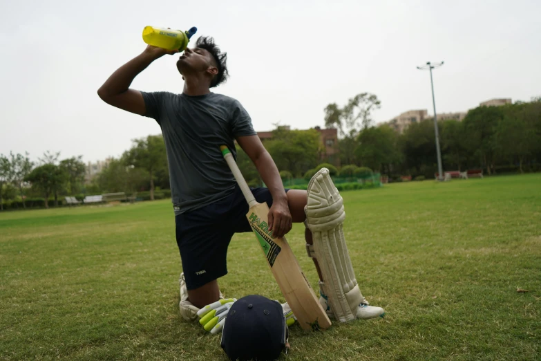 the man is drinking from a bottle while holding his cricket helmet on his knees