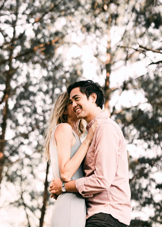 a smiling couple hugs in front of many trees