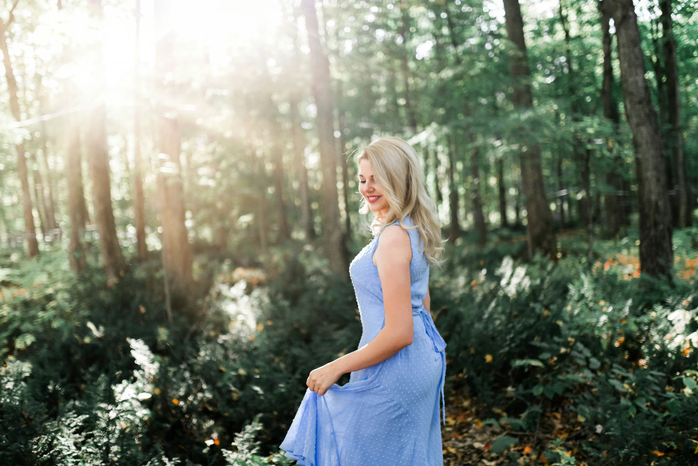 a pregnant woman in blue dress stands on the ground at the edge of a forest