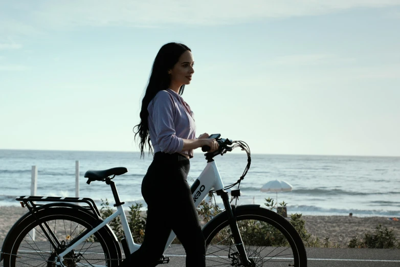 woman in white shirt and black pants walking her bicycle by the beach