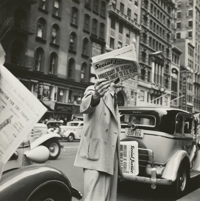 the man reads a newspaper near a classic car on a busy street