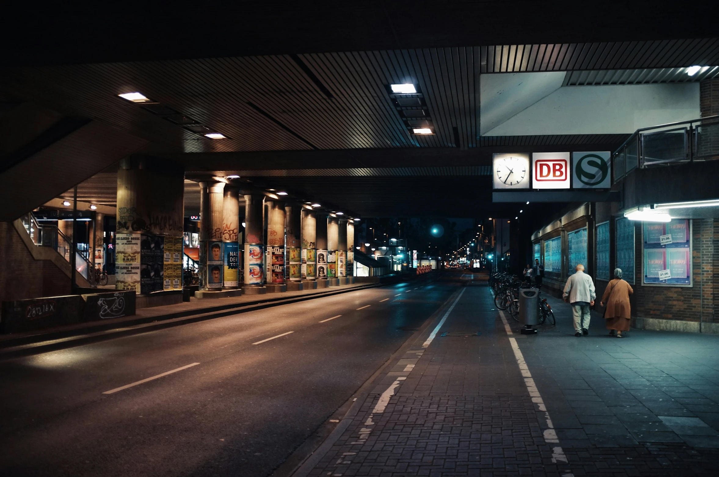 an empty city street at night with people walking by and the sidewalk is brick and there are many signs on the wall