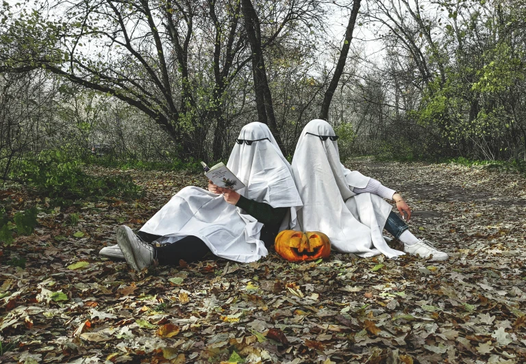 three women dressed as ghostes sit in a field with halloween pumpkins