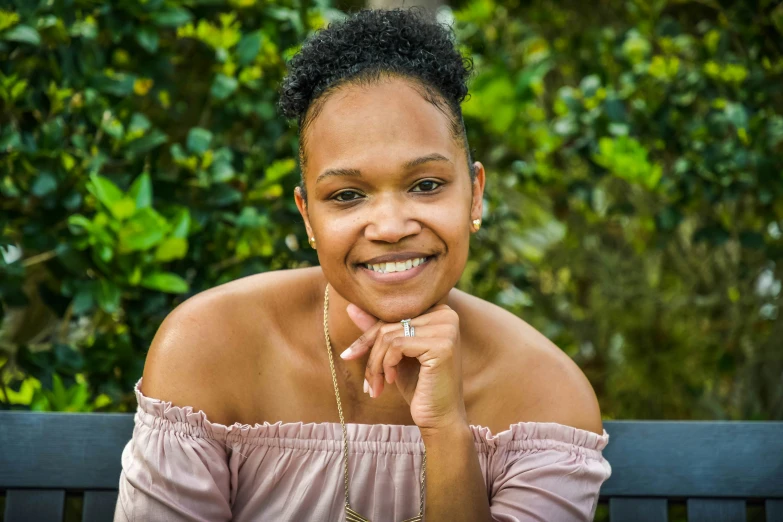 a black woman with short hair and piercings smiles as she sits on a bench in front of bushes