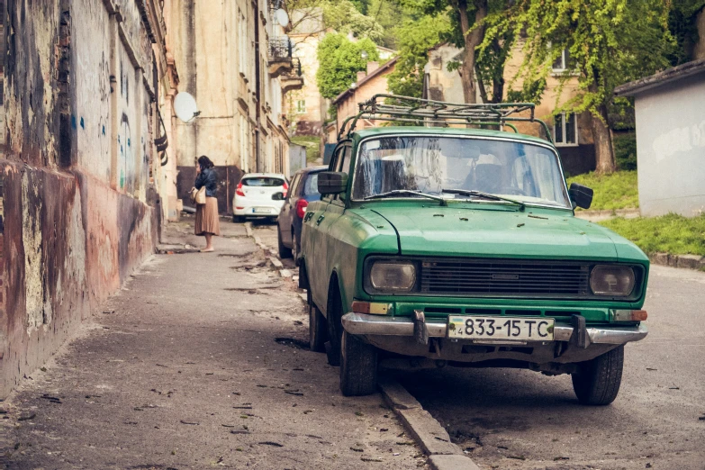 an old green truck driving down a street near a brick building