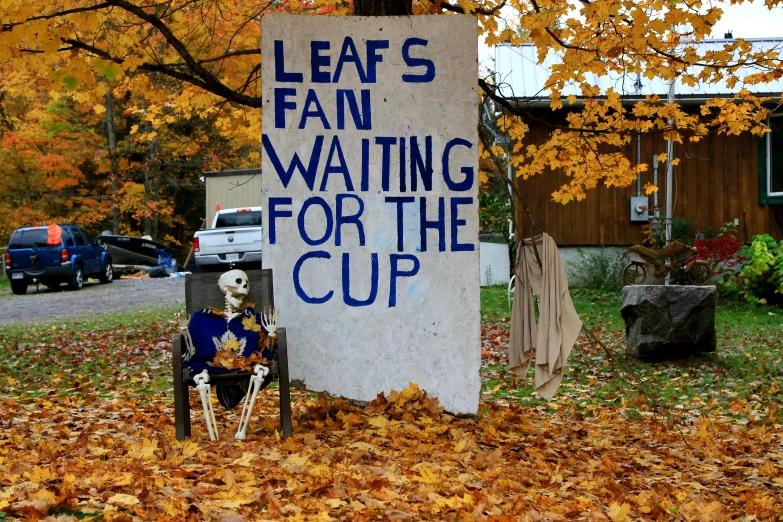 a halloween yard sign sits in leaves while a skeleton sits in a chair