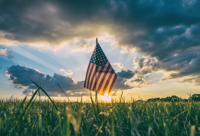 the sun peeking through the clouds is making a large american flag waving in the green grass