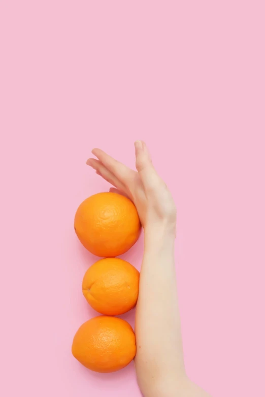 a female hand holding three oranges against a pink background
