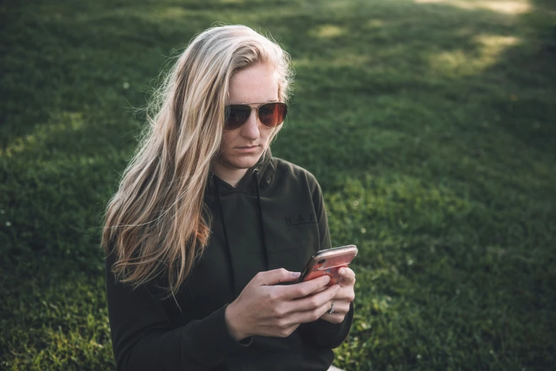 a young blond girl is sitting in a field while looking at her cell phone