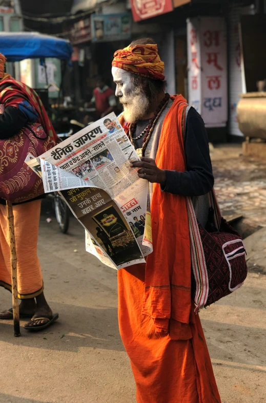 man with long gray and orange face paint on walking down street reading newspaper